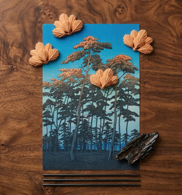 A rectangular art print or card featuring a stylized forest scene in blue tones with layered tree silhouettes, arranged on a dark wood surface. Several beige paper flower-like embellishments are placed around it, along with a dark bark piece and thin sticks at the bottom.