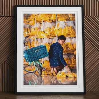 Framed photograph of a person handling produce in a market stall filled with yellow fruits or vegetables.