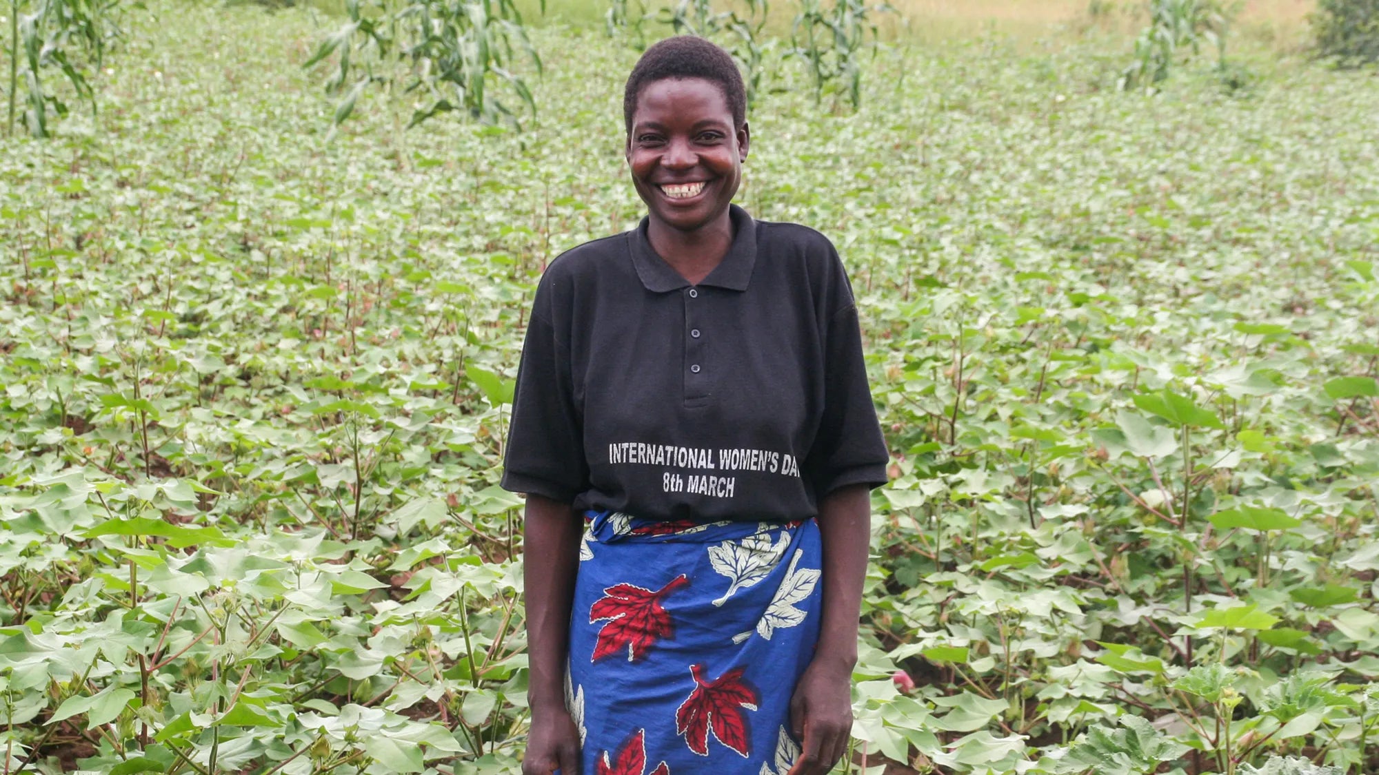 Someone wearing a black t-shirt and blue floral-patterned skirt stands with a bright smile