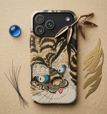 A smartphone case featuring a stylized tiger face illustration in black, beige, and blue, styled on a textured beige background with decorative props including a blue marble, a branch, and gold leaf.
