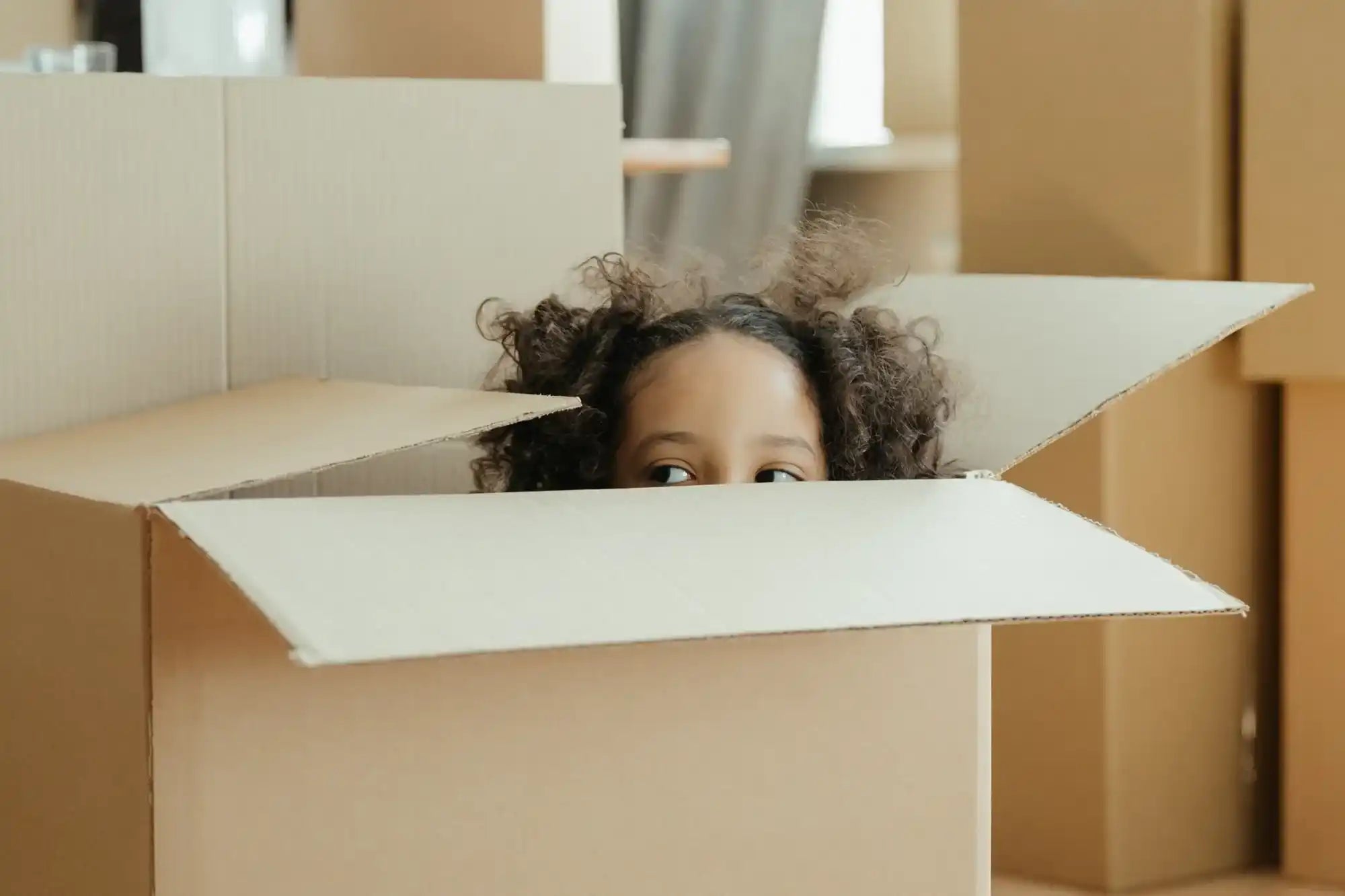 Cardboard moving box with curly hair and eyes peeking out from inside.