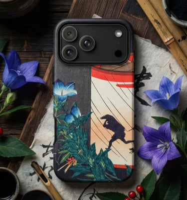 A phone case featuring a Japanese ukiyo-e style print with a frog, red lantern, and blue flowers, shown on a smartphone. The case is styled in a flat-lay setting with purple flowers, green leaves, and traditional wooden props.