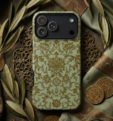 A phone case with an ornate gold damask pattern on a sage green background, shown on a dark phone. The case is styled in a flat-lay setting with olive leaves, bronze coins, and decorative metal filigree.