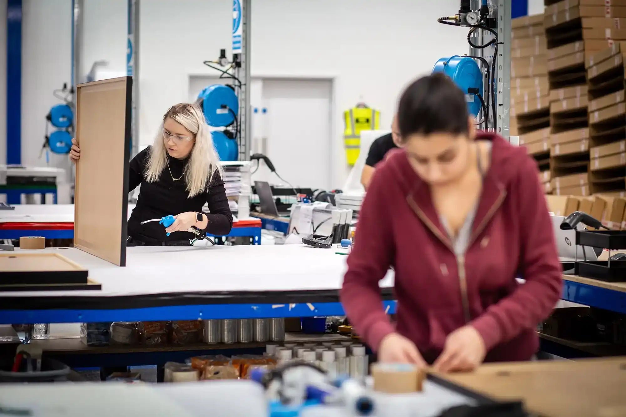 Two workers assembling or inspecting products at workstations with blue balloons visible in the background.